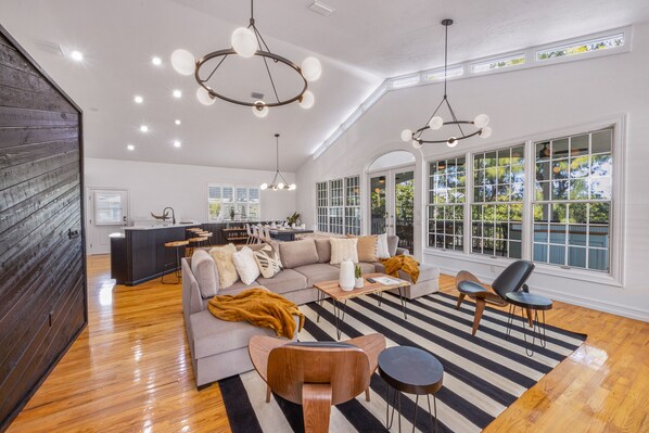 A spacious living room with a large sofa, a coffee table, and a striped rug. There are two pendant lights hanging from the ceiling and a large window with a view of trees.