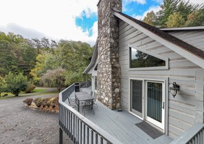 A house with a stone chimney, a covered deck with outdoor furniture, and a large window.