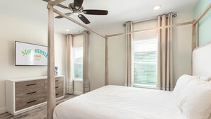 Une chambre à coucher avec un lit, un ventilateur de plafond, un téléviseur fixé au mur et une fenêtre avec des rideaux.