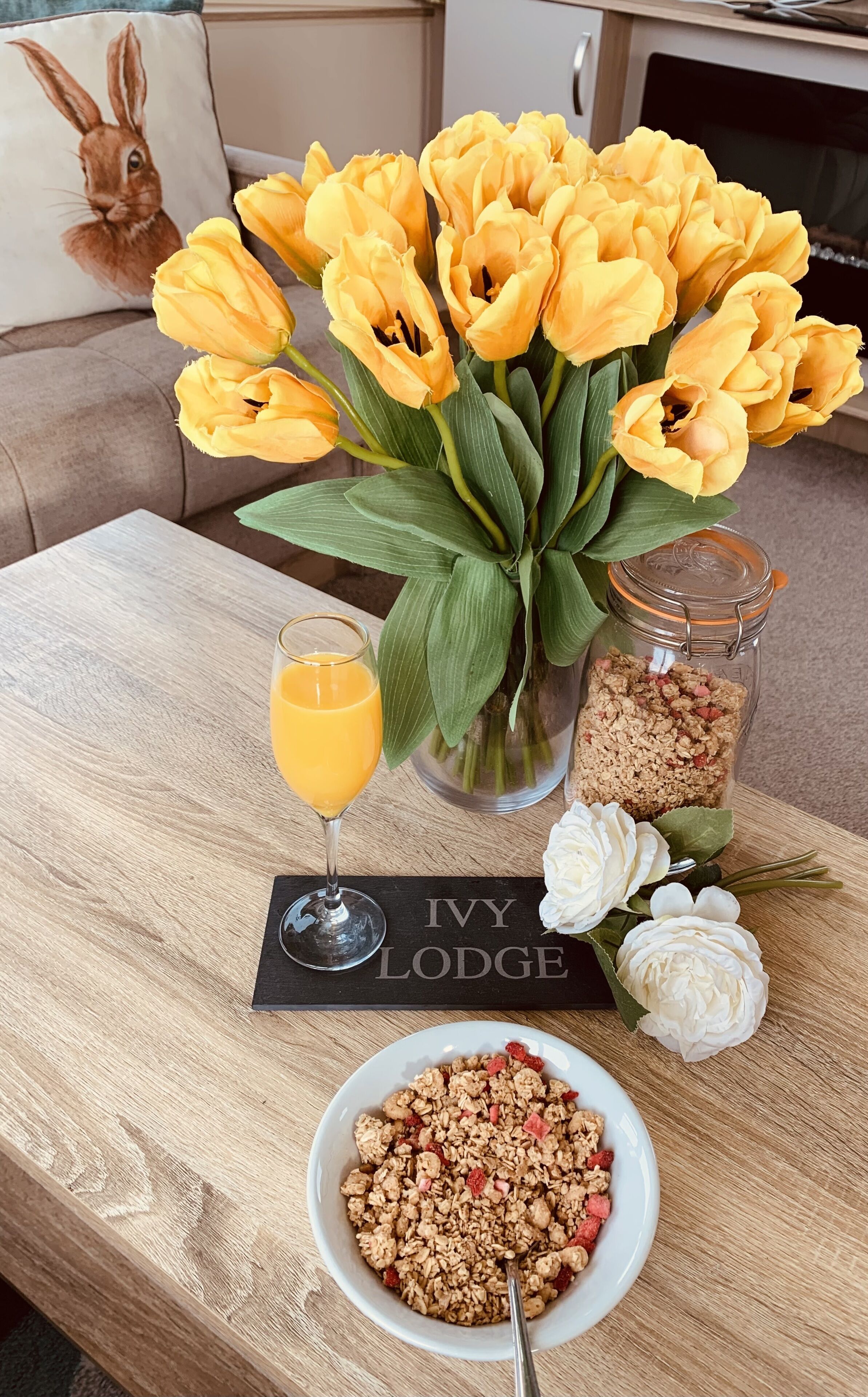 Une table en bois sur laquelle se trouvent un verre de jus d’orange, un bol de granola aux fraises, un vase de tulipes jaunes et une pancarte indiquant « IVY LODGE ».