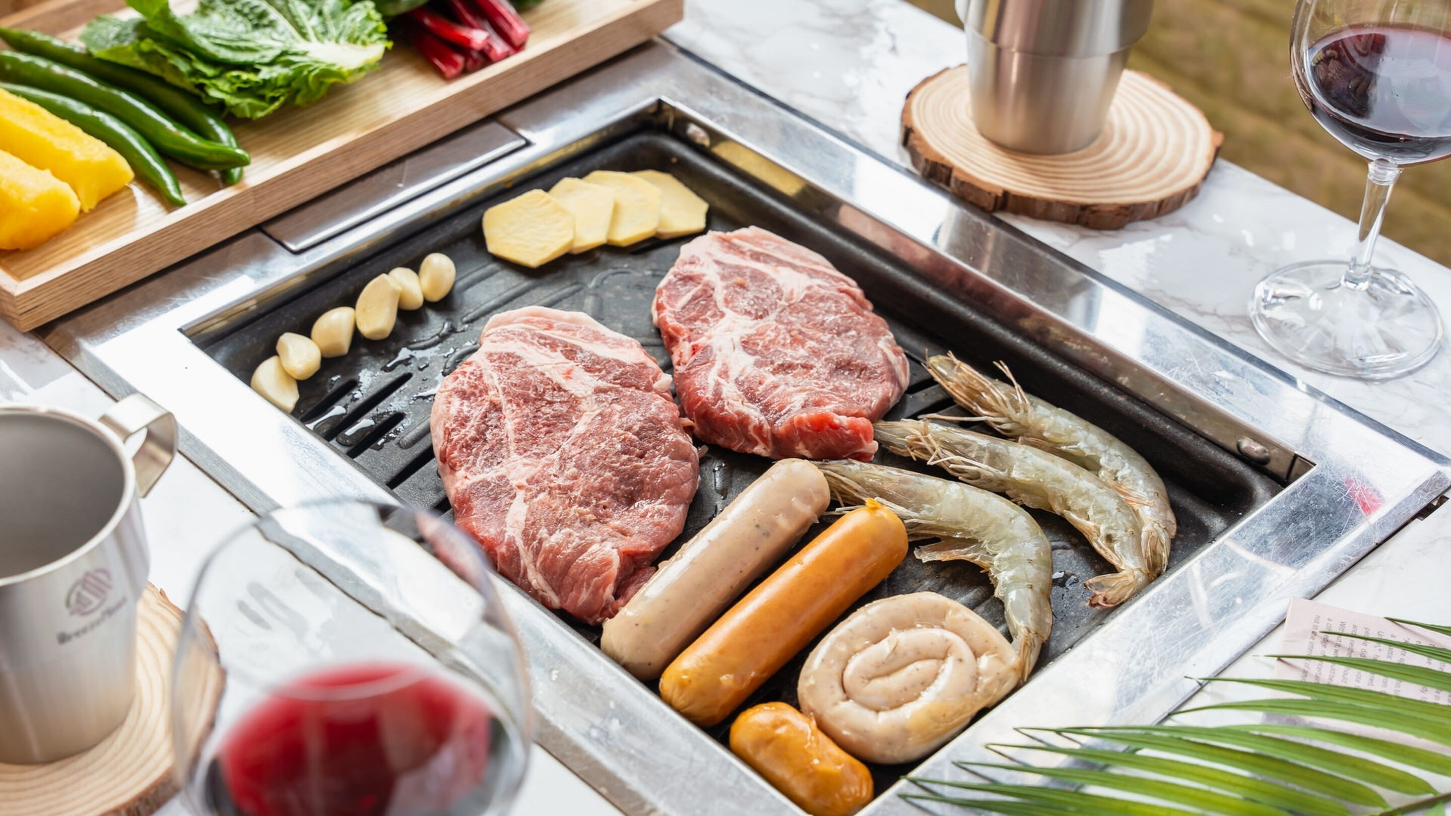 A grill with various meats and vegetables, a wine glass, and a wooden cutting board with fresh produce.