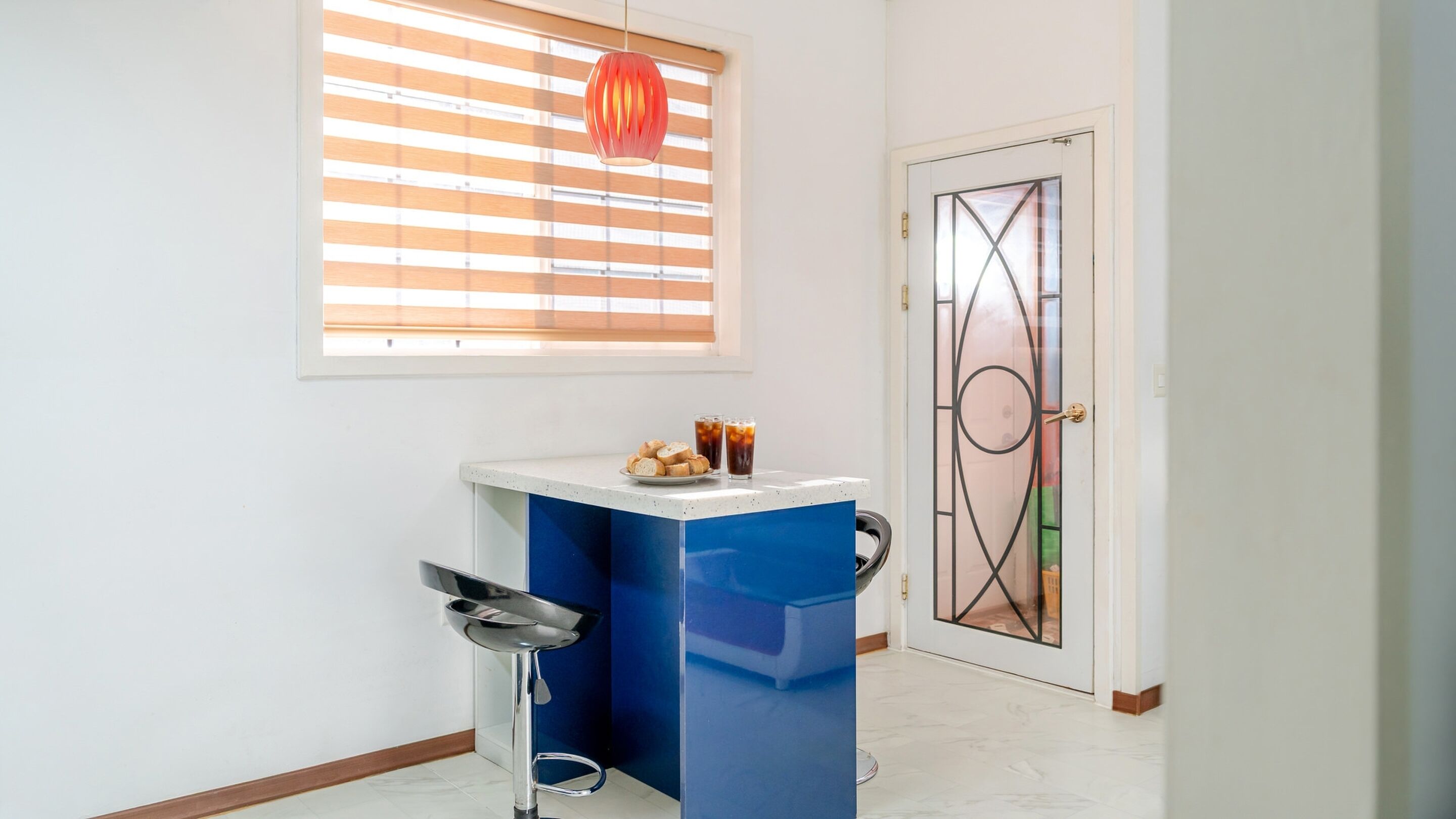 Una habitación con una puerta blanca, un cubo de basura azul, una pequeña mesa con una fuente de pan y dos vasos de té helado, y una ventana con persianas.