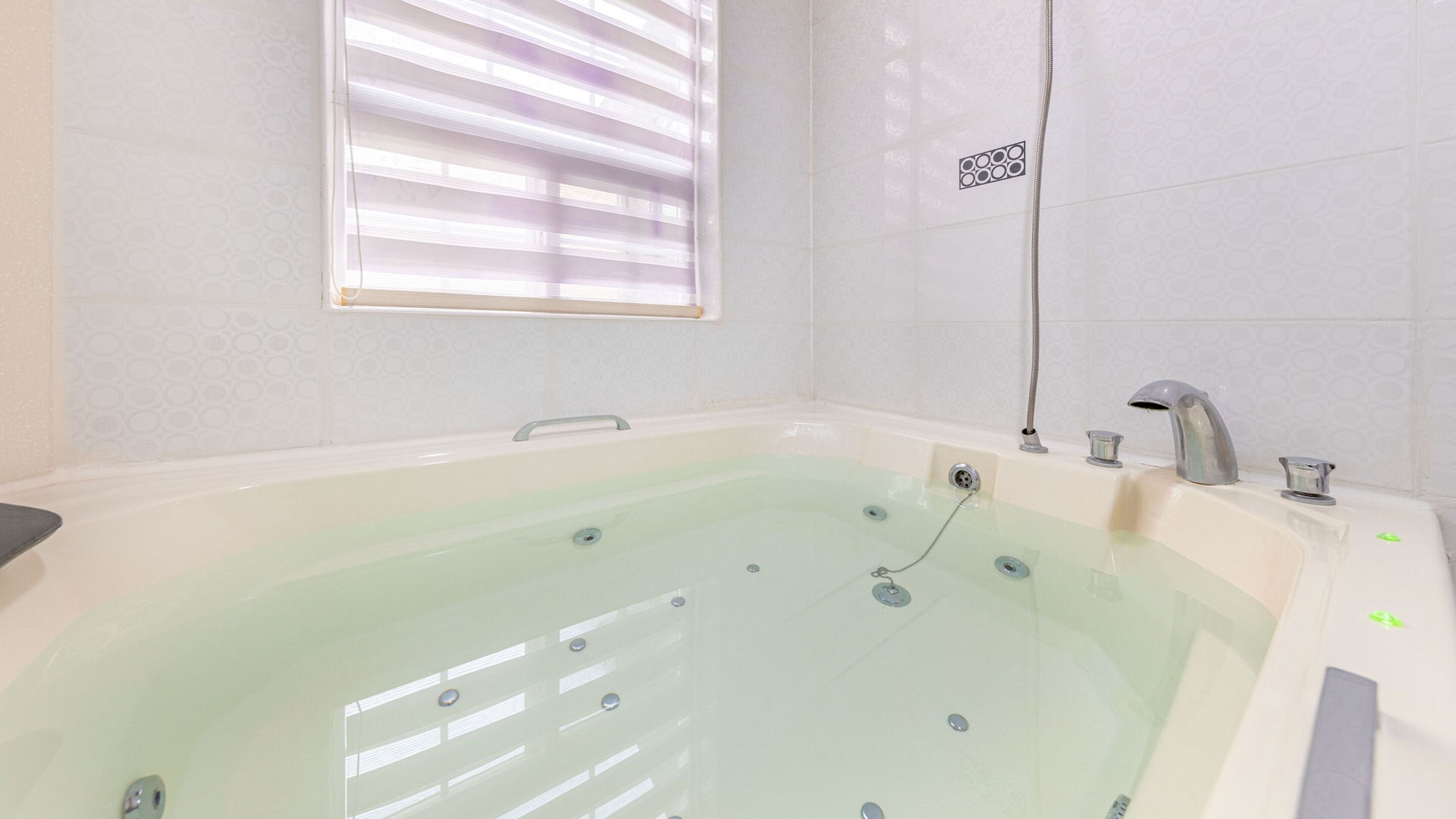 Une grande baignoire spacieuse, dotée d’un panneau de commande, d’une fenêtre avec stores et d’une pomme de douche.