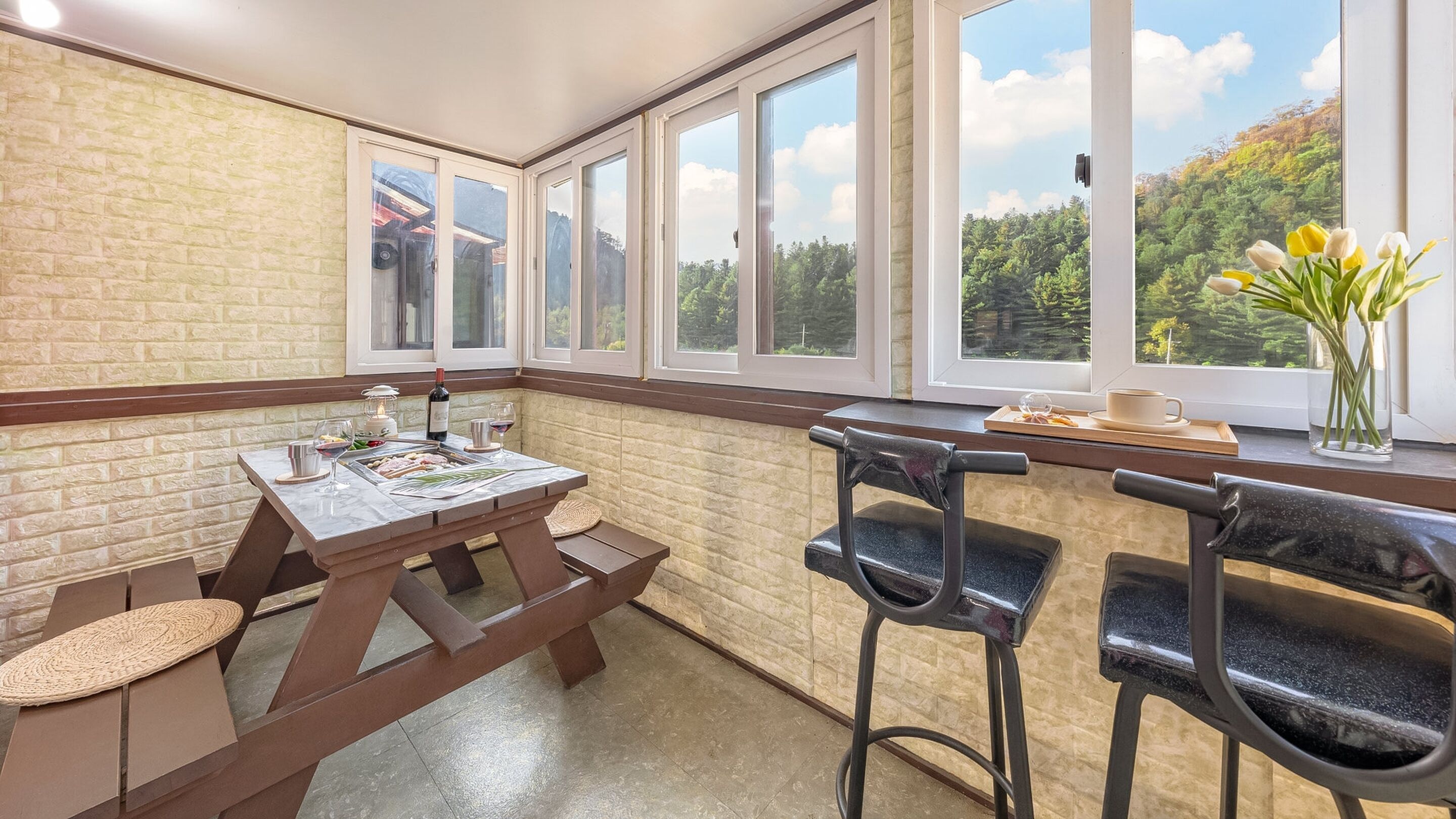 A dining area with a wooden table and chairs, a vase of flowers, and a view of trees through large windows.