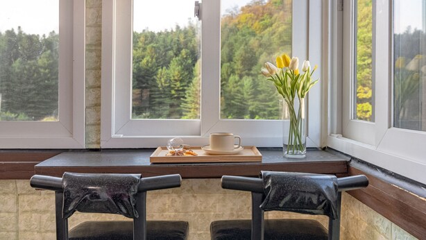 Un rincón acogedor con dos sillones, una taza, una bandeja con pasteles y un florero con flores, todo junto a una ventana con vistas a un bosque.