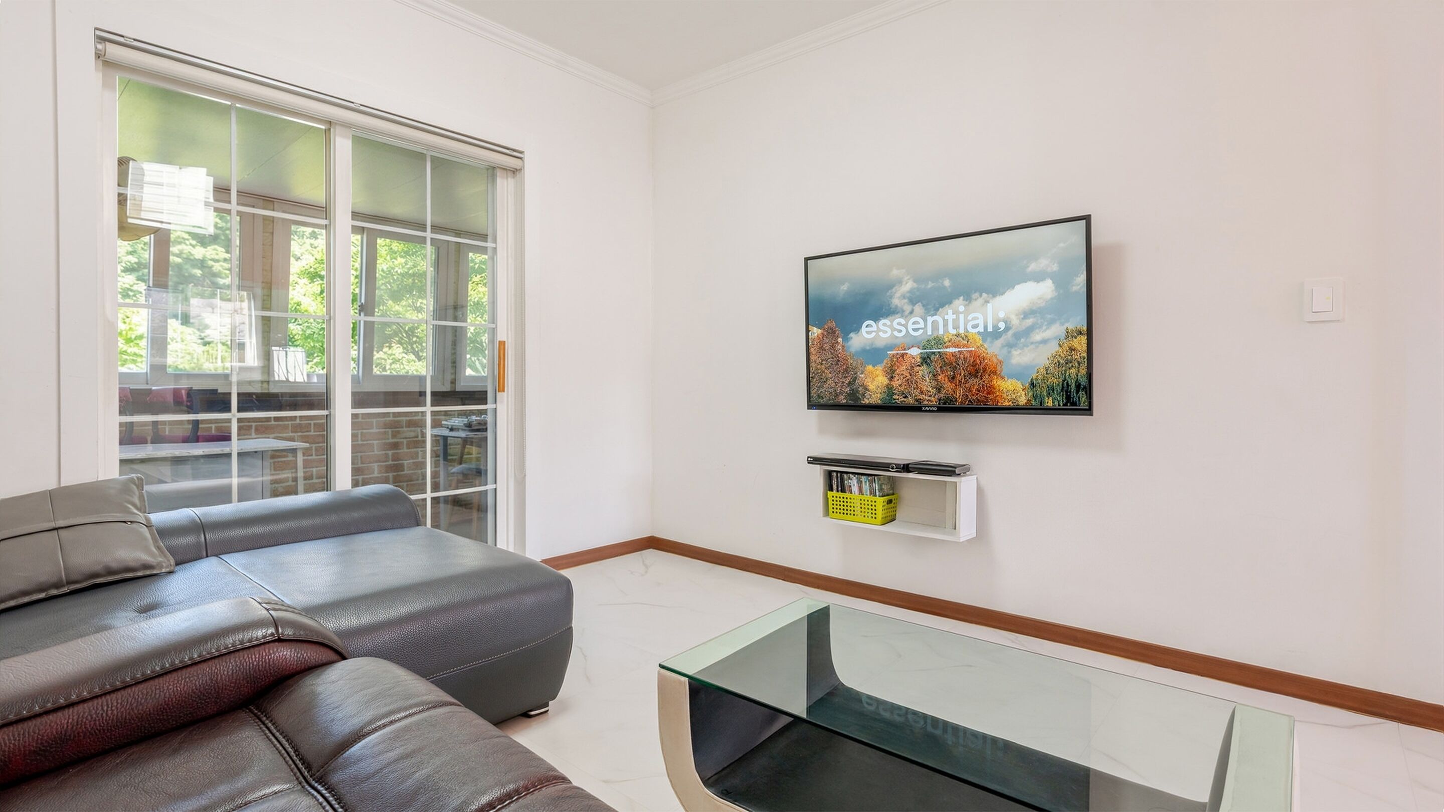 A modern living room with a leather sofa, a glass coffee table, a flat-screen TV mounted on the wall, and a sliding glass door leading to an outdoor area.
