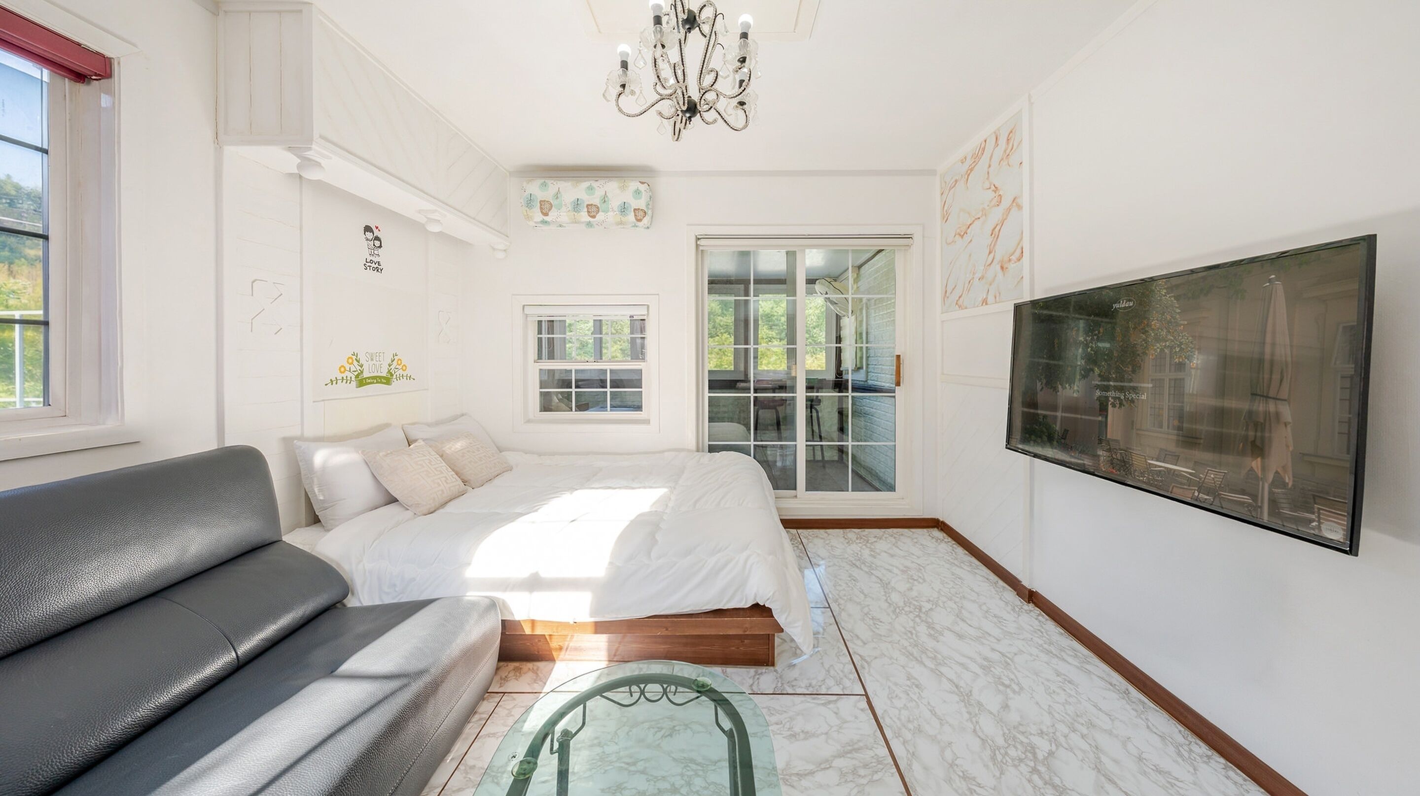 Une chambre à coucher moderne avec un lit, un canapé gris, une table basse en verre et un grand téléviseur fixé au mur.