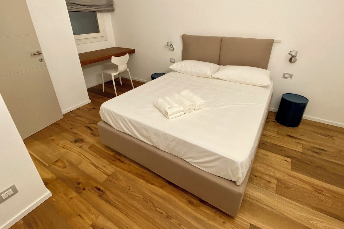 Un lit bien fait, avec du linge de lit blanc et des serviettes pliées, un petit bureau avec une chaise et une fenêtre avec un store gris.