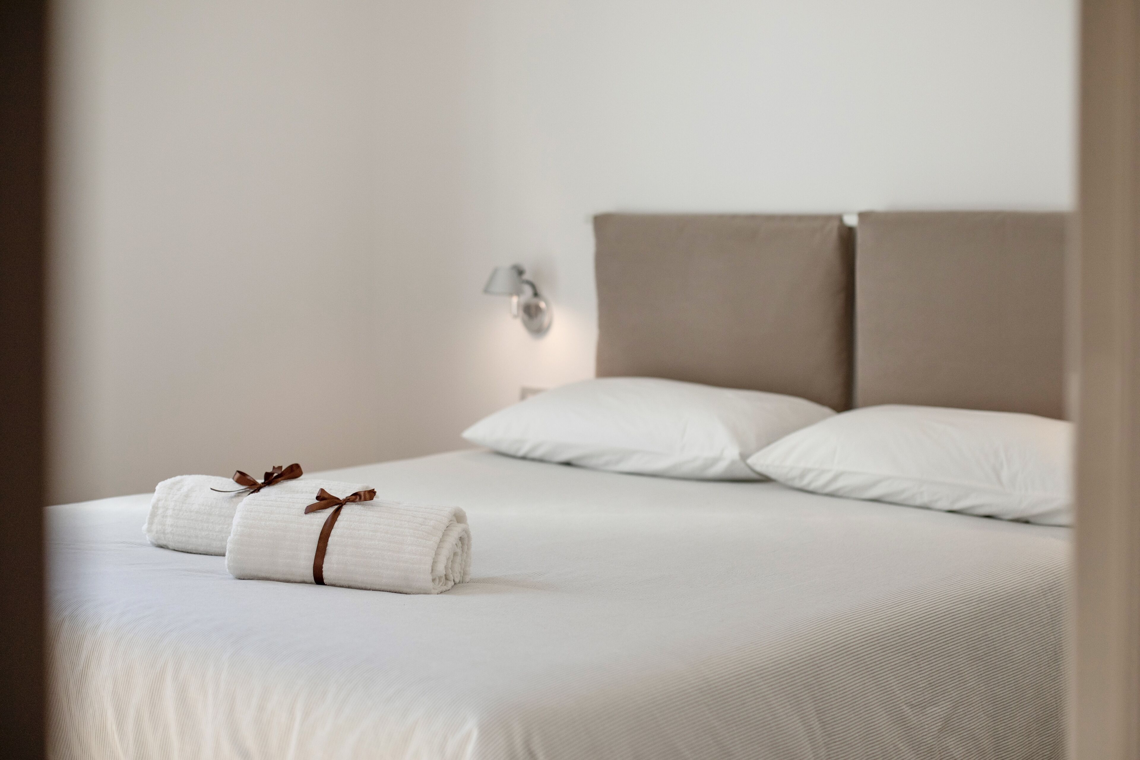 Een opgemaakt bed met witte lakens en twee opgerolde handdoeken met bruine linten, een beige hoofdbord, en een wandlamp.