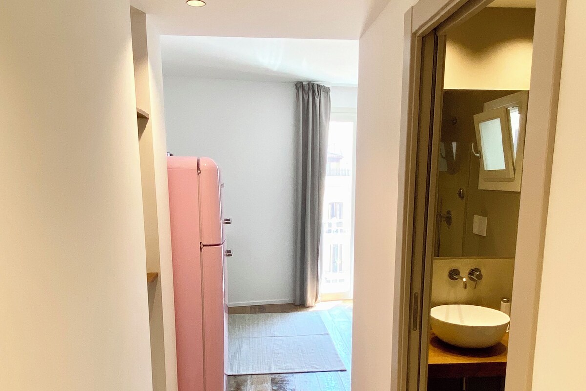 A narrow hallway with wooden flooring, a pink refrigerator, and a bathroom with a sink and mirror.