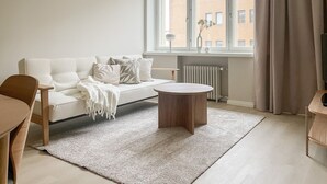 A modern living room with a sofa, a round coffee table, and a view of buildings through a large window. - Central 1BR next to the park (Helsinki)