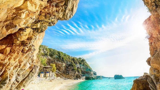 A coastal beach with clear turquoise waters, a rocky cliff, and a small building near the shore.