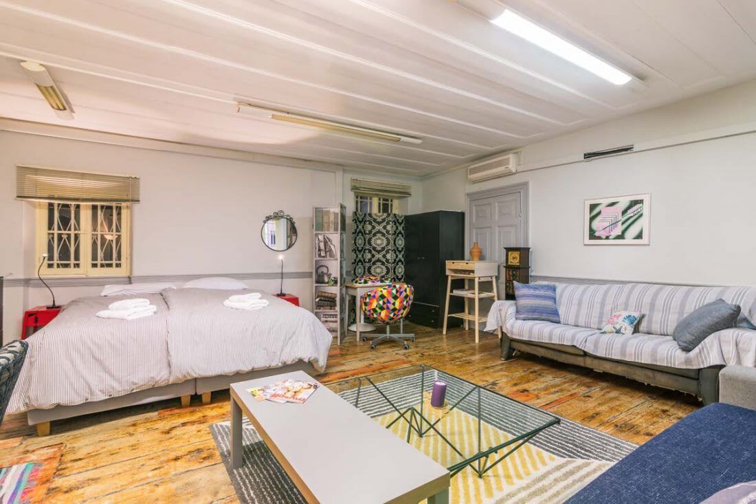Een kamer met twee bedden, een bank, een tafeltje en een stoel. Er is een raam met zonwering, een spiegel en een lijst met een afbeelding aan de muur.