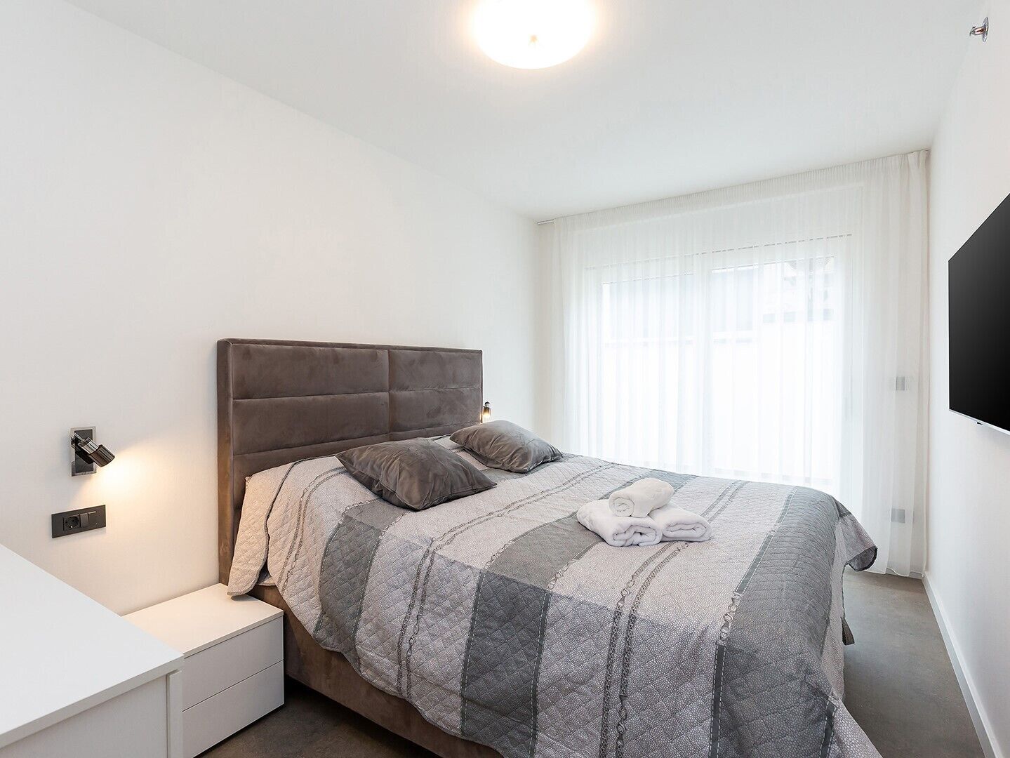 A neatly made bed with a quilted blanket, a bedside table with a lamp, and a window with sheer curtains.
