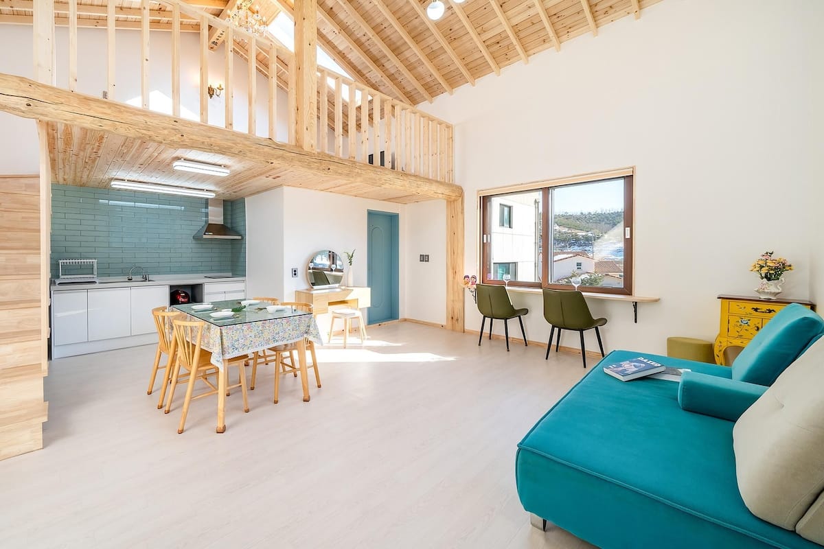 A modern interior with a staircase leading to an upper level, a dining area with a table and chairs, a sofa, and a window with a view.