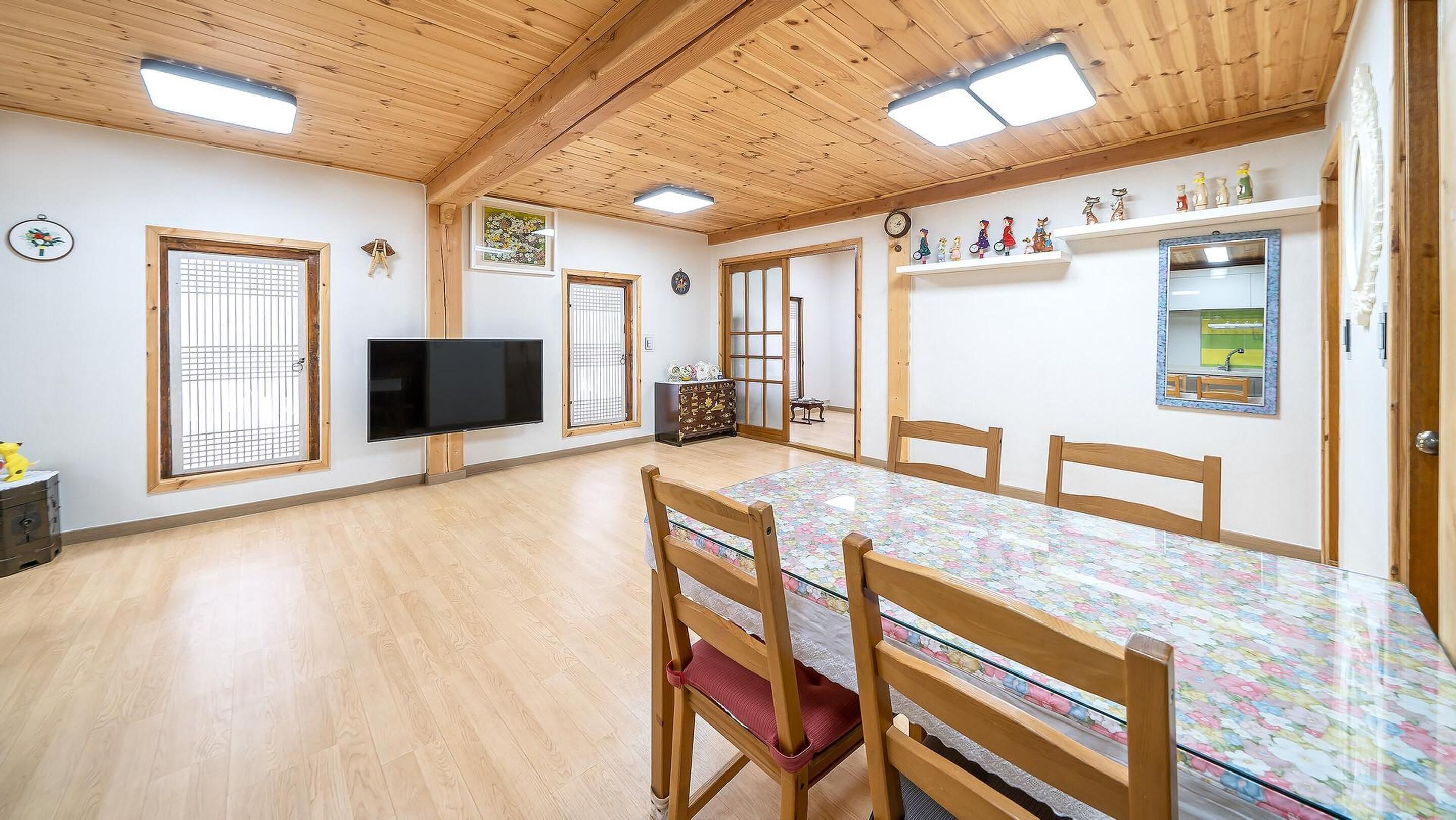 A spacious room with a wooden ceiling, a large flat-screen TV, a dining table with chairs, and a kitchen area visible through a glass door.