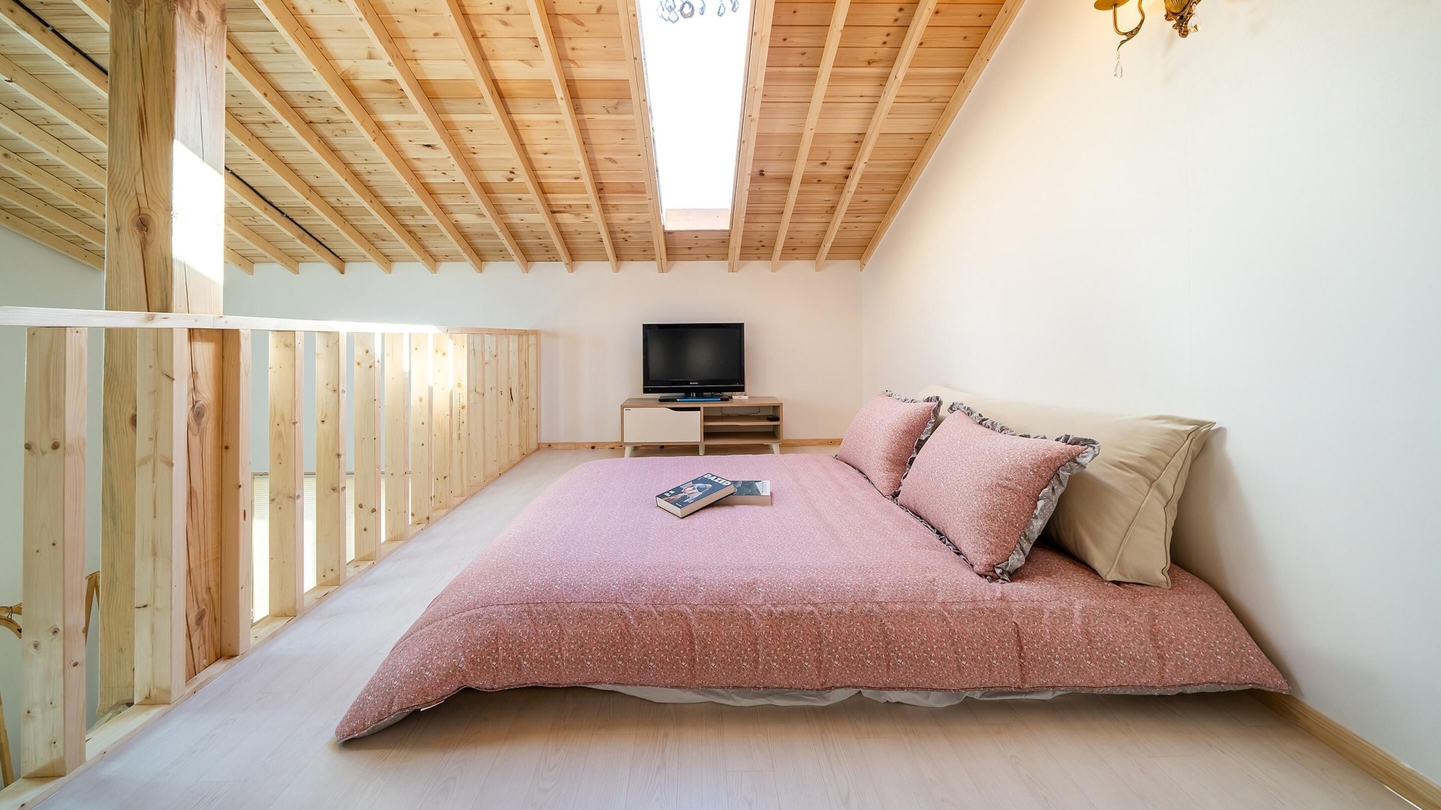 A bedroom with a sloped ceiling, a bed with pink bedding, a television on a wooden stand, and a window allowing natural light.