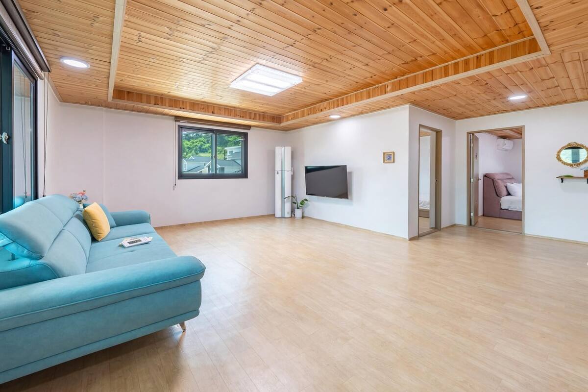A spacious living area with a blue sofa, a flat-screen TV, wooden flooring, and a ceiling with wooden beams and skylights.