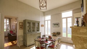 A dining area with a table set for four, a cabinet with glass doors, and a view of the outdoors through large windows. - Château Tournefeuille in the heart of the vineyards! (Néac)