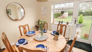 A round wooden dining table with blue plates, a vase with flowers, and a black pitcher. There are six wooden chairs around the table. A window with a view of a garden is visible in the background. - Glynderi (Cardigan)