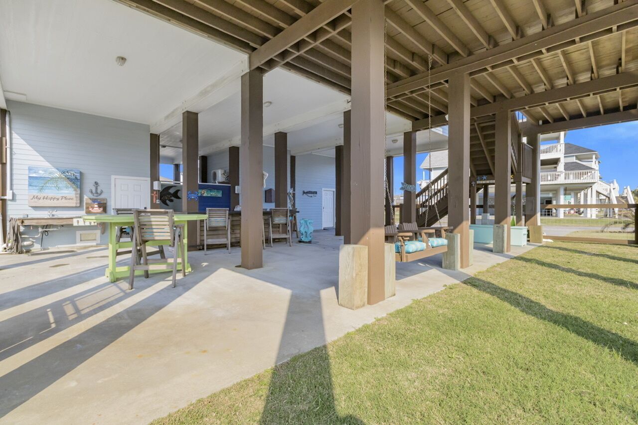 An open-air covered patio with concrete flooring, wooden pillars, and a view of a large house in the background.