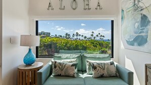 Une pièce avec vue sur les palmiers et l’océan, un canapé agrémenté de coussins à motifs, un vase bleu et un panneau mural sur lequel on peut lire « ALOHA ».