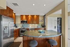A modern kitchen with wooden cabinets, stainless steel appliances, a granite countertop, and a built-in island with bar stools. - Wailea Elua, #2003 1 Bedroom Condo by RedAwning (Kihei)
