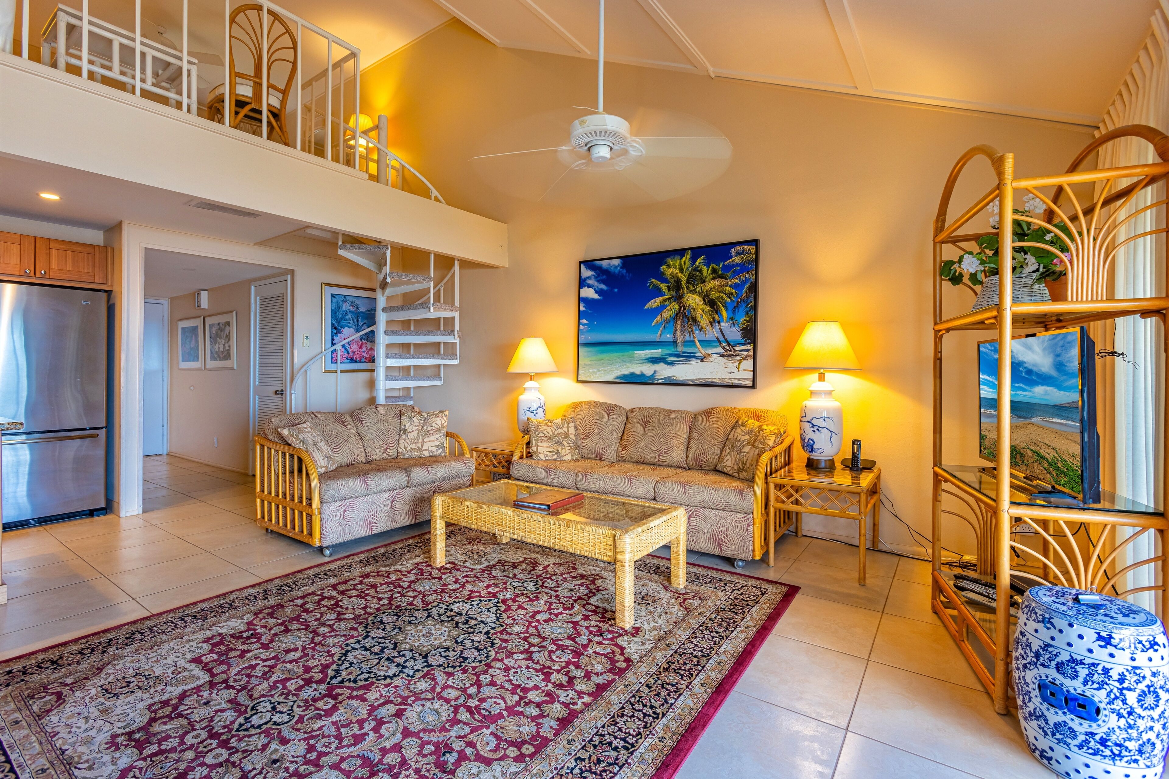 A spacious living room with a large sofa set, a coffee table, and a decorative rug. There is a staircase leading to an upper level, and a large window with a view of the beach.