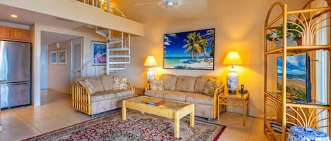 A spacious living room with a large sofa set, a coffee table, and a decorative rug. There is a staircase leading to an upper level, and a large window with a view of the beach.