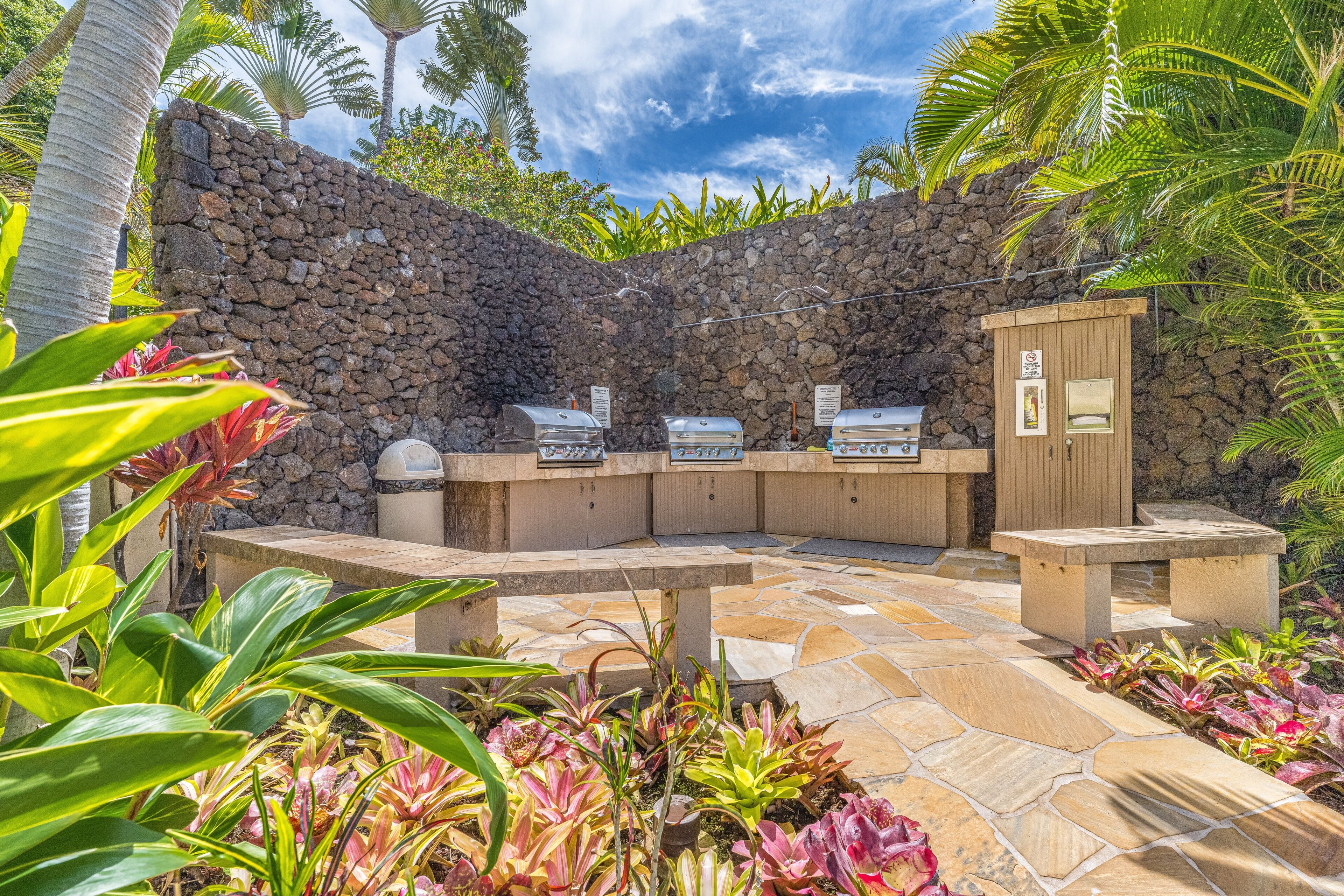 An outdoor barbecue area with stone walls, a grill station, and a seating area surrounded by tropical plants.