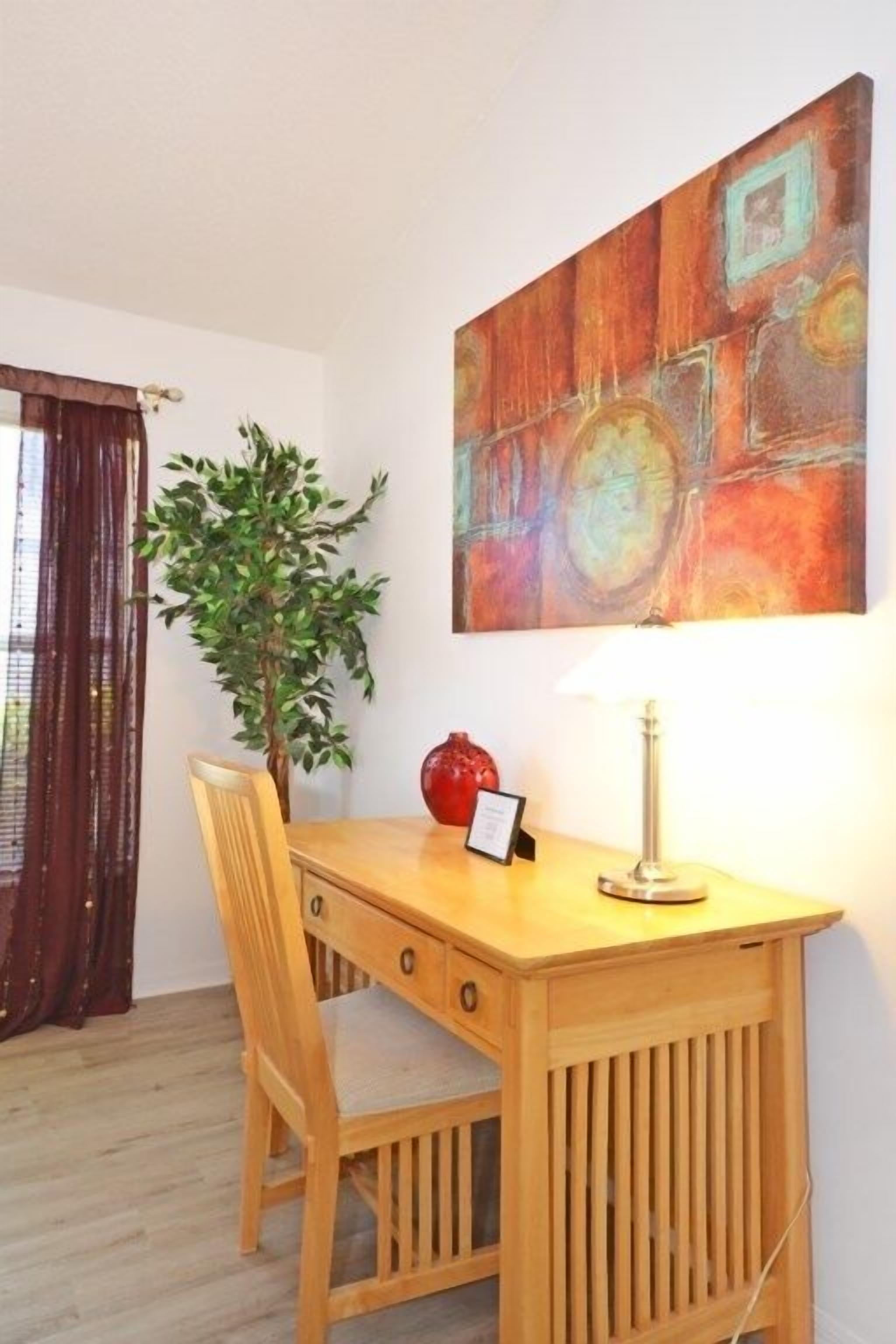 A wooden desk with a lamp, a chair, a potted plant, and a colorful abstract painting on the wall.