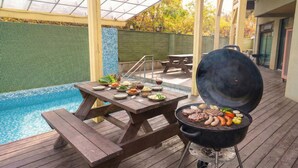 Una terraza de madera con una parrilla, piscina y mesa de comedor con comida y bebidas.