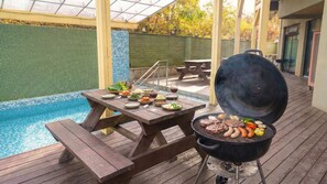 Una terraza de madera con una parrilla, piscina y mesa de comedor con comida y bebidas.