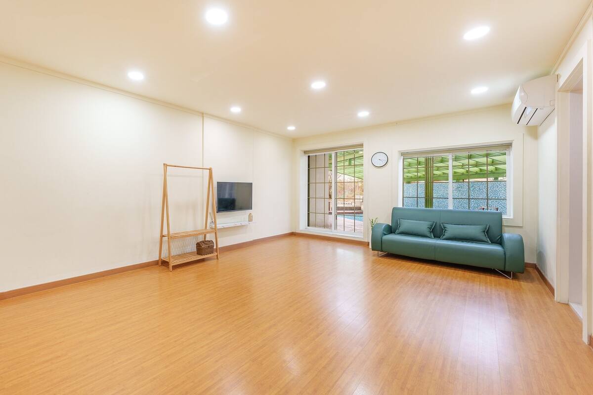A spacious room with a wooden floor, a green sofa, a TV on a stand, and a large window with a view of greenery.