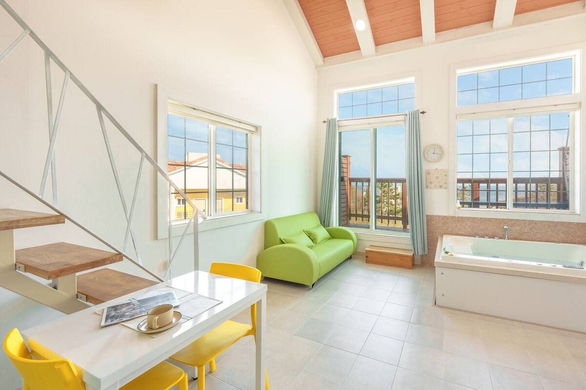 A bright room with a green sofa, a white bathtub, and a dining area with yellow chairs.