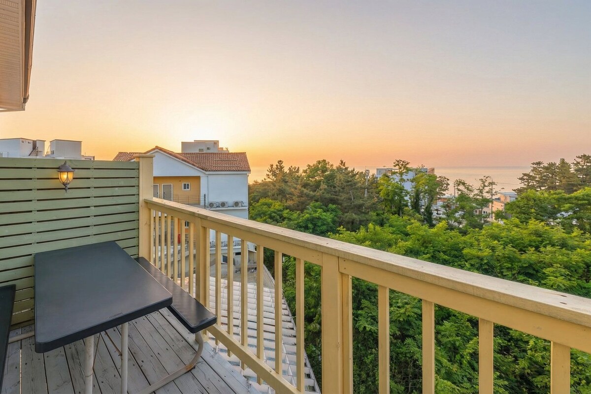 A balcony with a table and chairs, overlooking a residential area and the sea during sunset.