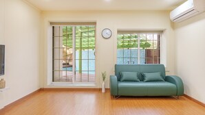 A living room with a green sofa, a clock on the wall, and a sliding glass door leading to an outdoor area with a pool. - Gangleung Laimbichipensyeon (Gangneung)