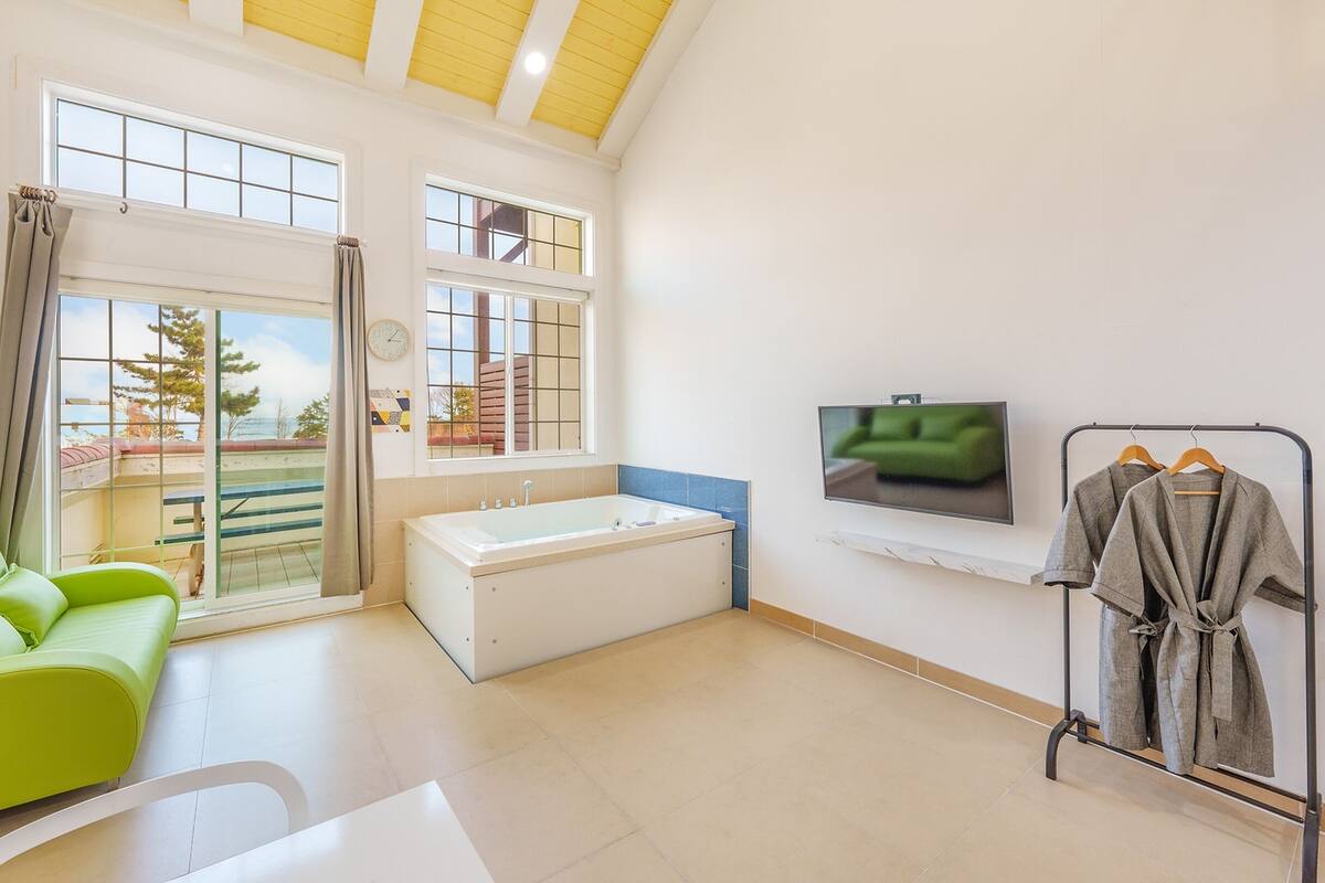 A bright room with a green sofa, a bathtub, a TV, and a clothing rack.