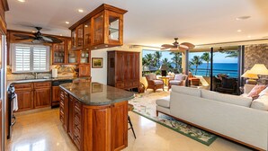 A spacious living area with a large window overlooking the ocean, a kitchen with wooden cabinetry, and a seating area with a sofa and chairs. - Polo Beach Club Two Bedrooms - Ground Floor by Coldwell Banker Island Vacations (Kihei)