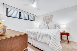 A bedroom with a bed, bedside tables, a ceiling fan, a window with blinds, and a wall-mounted macrame piece. - Kihei Surfside, #110^ 1 Bedroom Condo by RedAwning (Kihei)