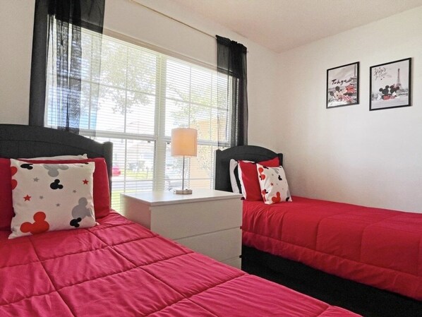 A bedroom with two beds, each with red bedding and Mickey Mouse themed pillows. There is a window with blinds, a bedside table with a lamp, and two framed pictures on the wall.