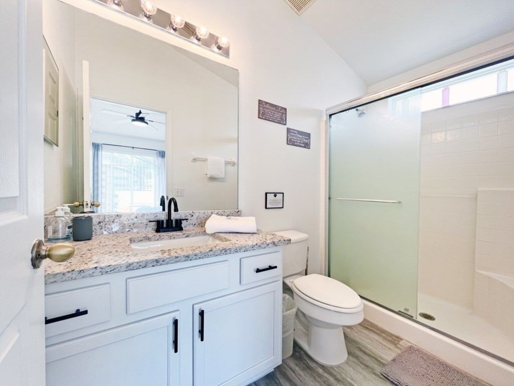 A modern bathroom with a granite countertop, white cabinets, a glass shower enclosure, and a visible window.