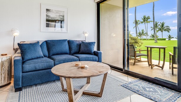 A modern living room with a blue sofa, a round wooden coffee table, and a large sliding glass door leading to an outdoor area with a view of palm trees and a golf course. - Kihei Surfside, #113 1 Bedroom Condo by RedAwning (Kihei)