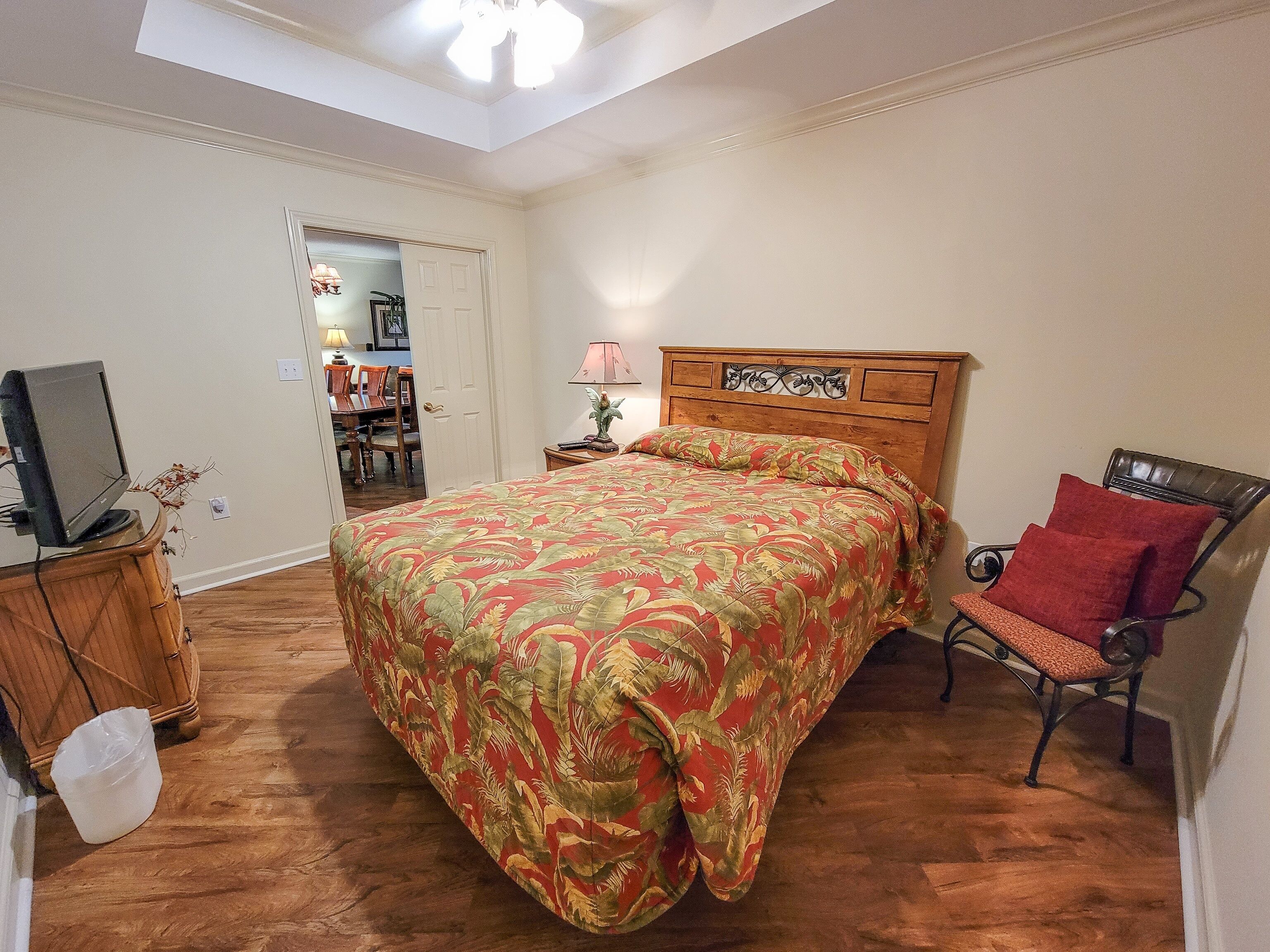 Une chambre avec un lit en bois, une chaise avec des coussins rouges, un téléviseur posé sur un meuble en bois, et une porte donnant sur un coin repas.