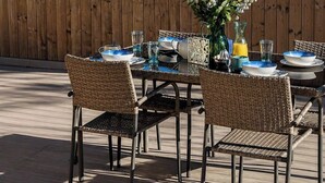 An outdoor dining area with a table set for four, surrounded by wicker chairs, a wooden fence, and a view of the ocean.