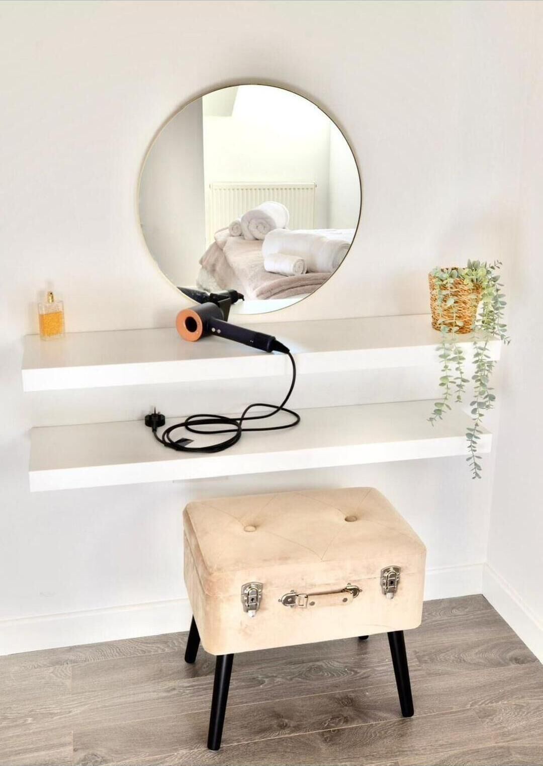 Un miroir rond accroché au mur, un sèche-cheveux posé sur une étagère, une petite plante et un tabouret de style vintage.
