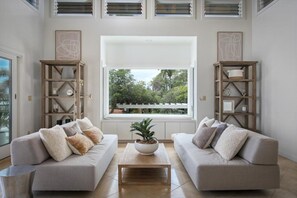 A modern living room with two sofas, a coffee table, and a large window with a view of greenery. - Puuwai Alii Estate: Stunning Luxury Home with Pool, Hot Tub & Ocean Views,AC! (Kailua Kona)