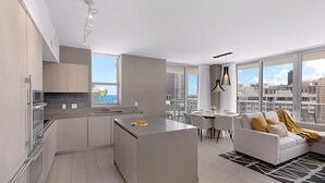 A modern apartment with a kitchen, dining area, and living room. It features a large island in the kitchen, a dining table with chairs, and a sofa with a view of the city. - Wrap Around Balcony|Insane Amenities|Amazing Pools (Hollywood)