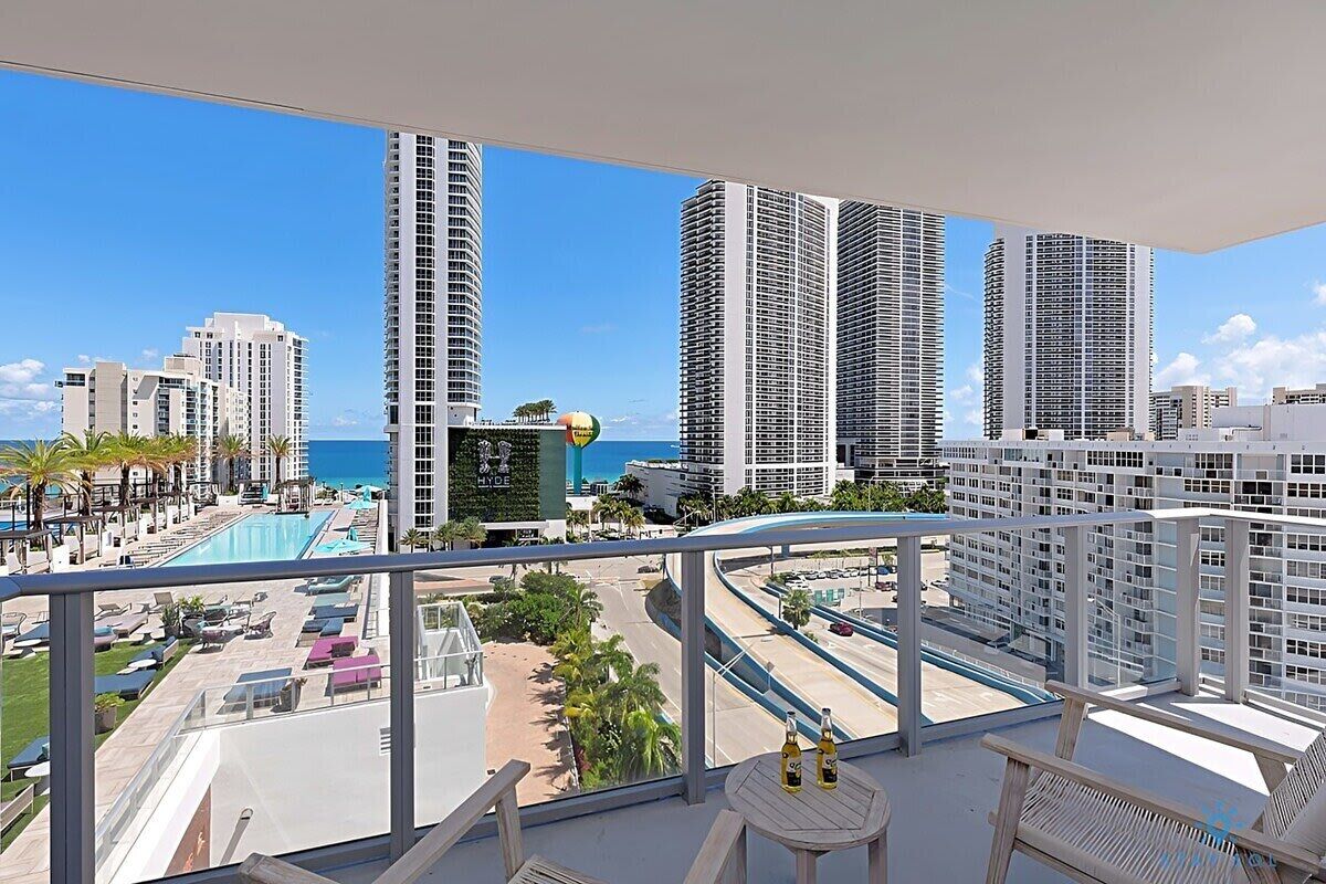 Ein Balkon mit Blick auf einen Hotelkomplex, einen Swimmingpool und einen wolkenlosen, blauen Himmel.