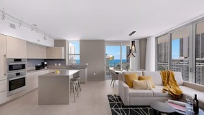 A modern apartment with a kitchen, dining area, and living room. It features a large window with a city view, a sofa with cushions, and a small table with a bottle and glasses. - Wrap Around Balcony|Insane Amenities|Amazing Pools (Hollywood)